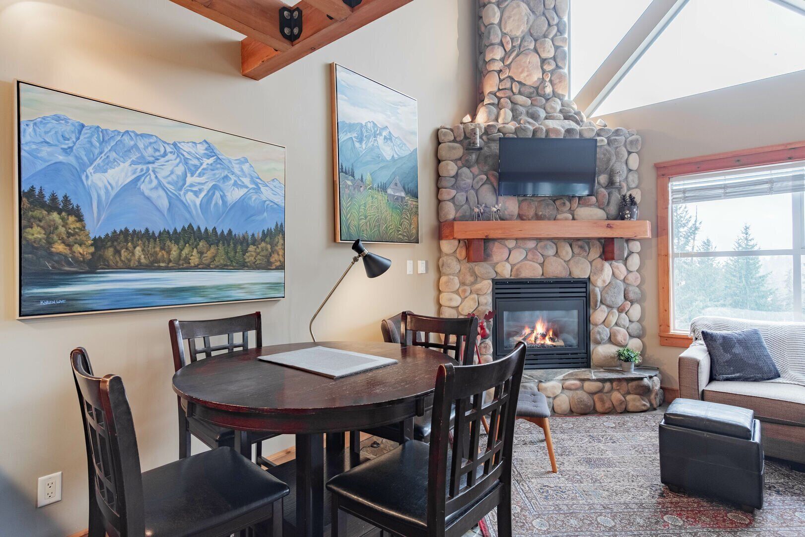 A cozy living room with a stone fireplace, a dining table with chairs, and two large paintings on the wall.