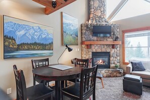 A cozy living room with a stone fireplace, a dining table with chairs, and two large paintings on the wall. - Large groups and families | Free parking | Hot tub (Whistler)