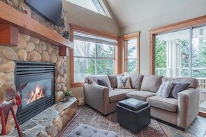 Ein gemütliches Wohnzimmer mit Kamin aus Stein, einer Eckcouch und großen Fenstern mit Blick ins Freie.