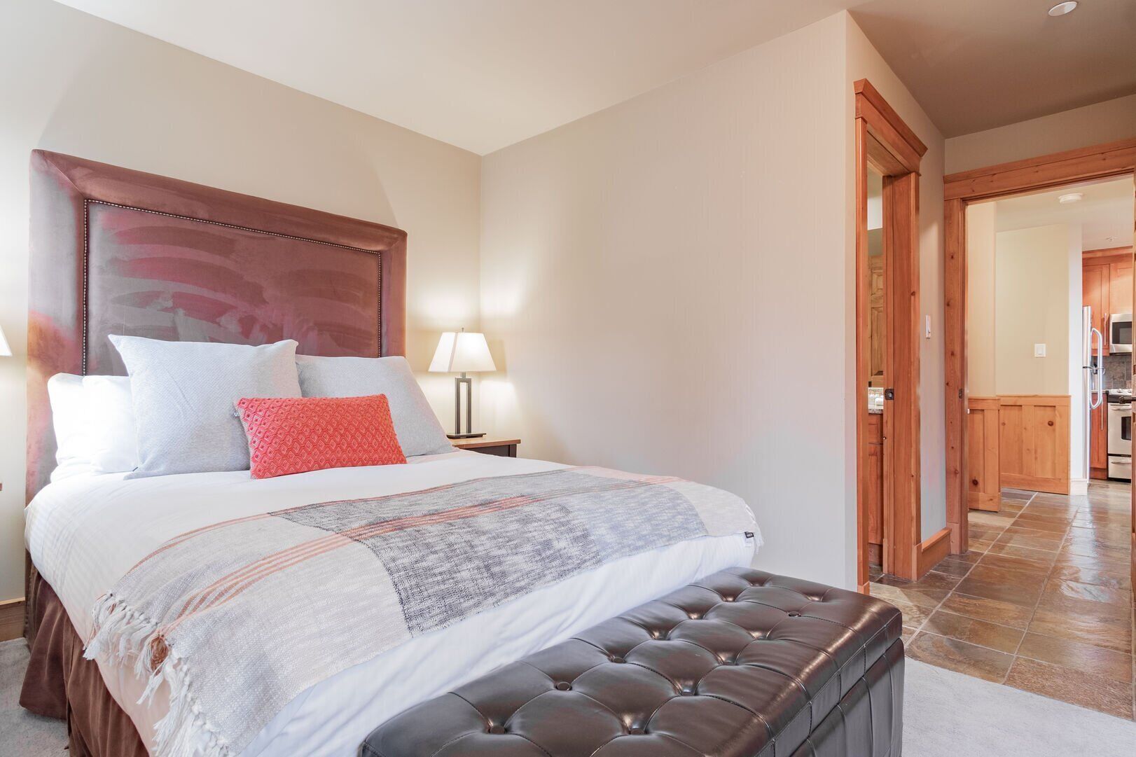 A bedroom with a large bed, a wooden headboard, a bedside table with a lamp, and a view into the kitchen.