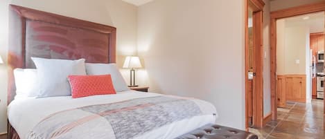 A bedroom with a large bed, a wooden headboard, a bedside table with a lamp, and a view into the kitchen.