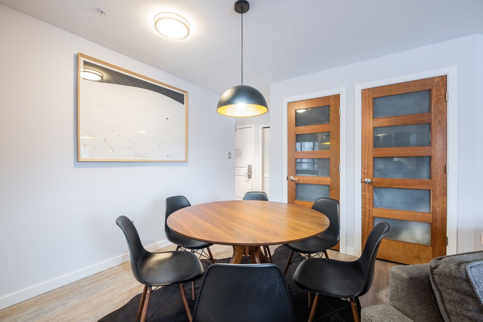 Un espace repas comprenant une table ronde en bois, des chaises noires, un luminaire suspendu et deux portes en bois dotées de panneaux de verre.