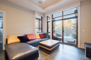 Ein modernes Wohnzimmer mit einem Ecksofa, einem Hocker und einer großen Schiebetür, die auf einen Balkon führt.
