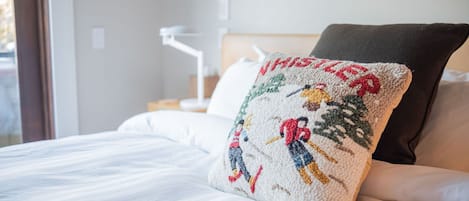 Una cama con ropa de cama blanca, un almohadón con diseño navideño y un almohadón de color oscuro.