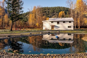 A white cabin with a pond in front, surrounded by autumn trees and a forested hill.