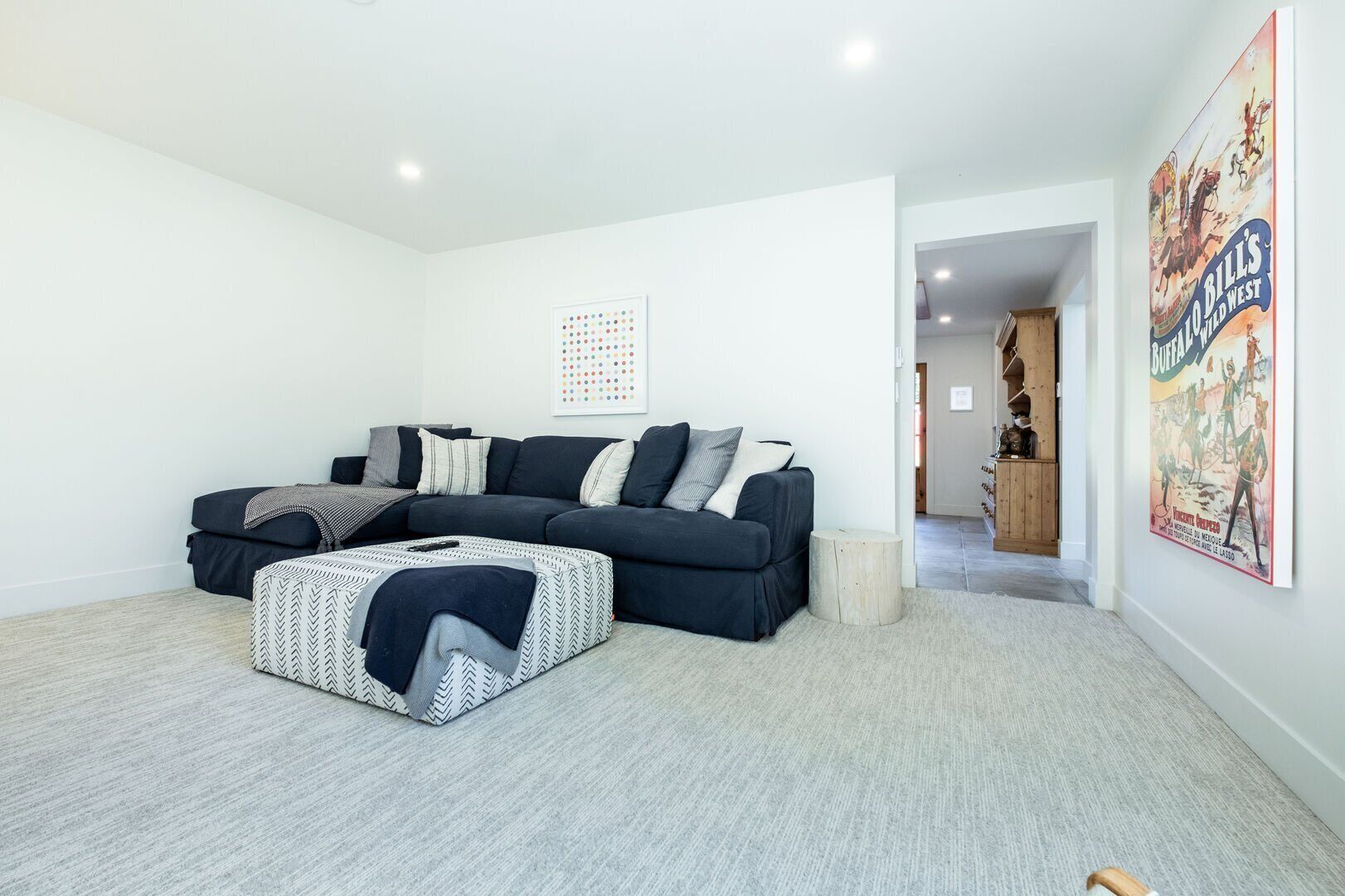 A living room with a sectional sofa, a patterned ottoman, and a poster on the wall.