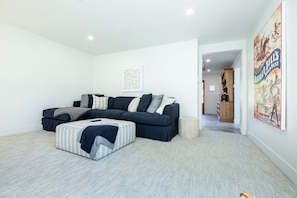 A modern living room with a sectional sofa, a patterned ottoman, and a poster on the wall.