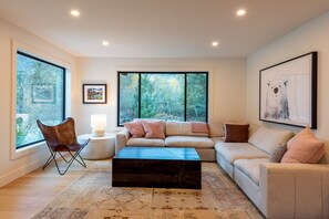 A modern living room with a sectional sofa, a coffee table, a side table with a lamp, and a chair. There are large windows with a view of trees. - Secluded house in Pemberton | Mount Currie Views | Sauna (Whistler)