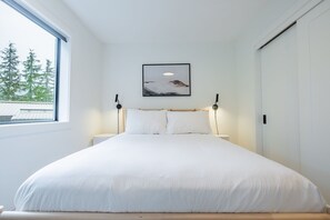 Ein Schlafzimmer mit einem großen Bett, einem Fenster mit Blick auf Bäume, einem gerahmten Bild an der Wand und einer Nachttischlampe.