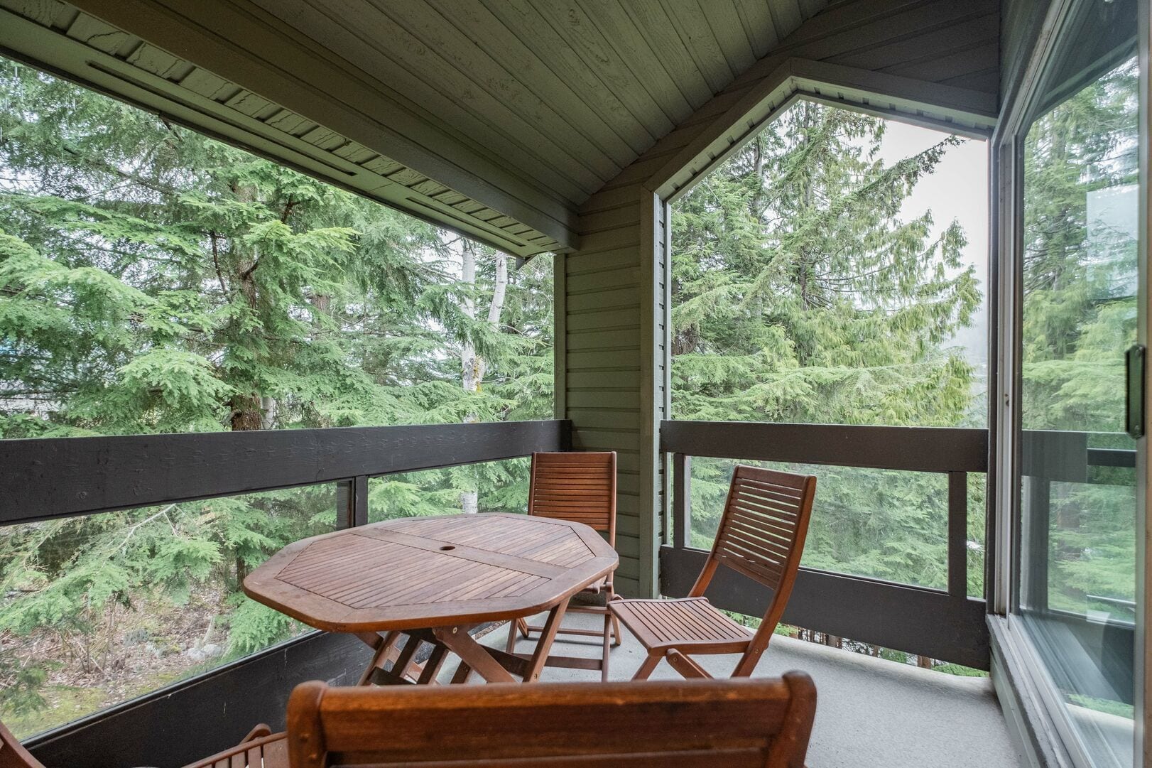 Un espace extérieur abrité, doté d’une table et de chaises, et entouré de murs de verre offrant une vue sur une forêt dense.