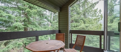 Un espace extérieur abrité, doté d’une table et de chaises, et entouré de murs de verre offrant une vue sur une forêt dense.