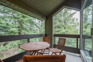 A covered outdoor area with a table and chairs, surrounded by glass walls offering a view of dense forest. - Gorgeous 2+Loft | Creekside | Free Parking (Whistler)