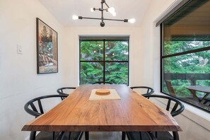 A dining area with a wooden table, black chairs, a hanging light fixture, a framed picture, and a large window with a view of trees. - Gorgeous 2+Loft | Creekside | Free Parking (Whistler)