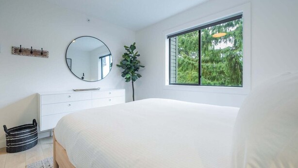 Ein Schlafzimmer mit einem Bett, einer Kommode, einem runden Spiegel, einer Topfpflanze und einem Fenster mit Blick auf Bäume.