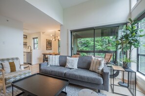 Ein modernes Wohnzimmer mit einem grauen Sofa, einem Couchtisch und Blick auf einen Balkon mit Pflanzen.