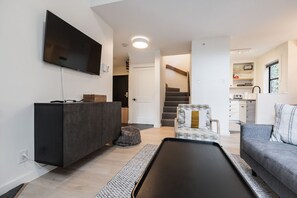 Un salon moderne avec un téléviseur à écran plat, un meuble en bois foncé, un canapé gris, un fauteuil à motifs et un escalier menant à la cuisine.
