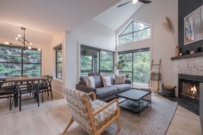 A spacious living room with a high ceiling, large windows, a fireplace, and a dining area with a table and chairs. - Gorgeous 2+Loft | Creekside | Free Parking (Whistler)