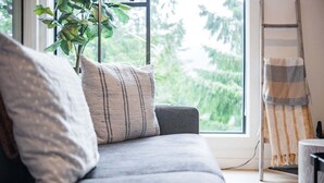 Ein graues Sofa mit gestreifter Dekokissen, eine Pflanze und eine Leiter vor einem Fenster mit Blick auf Grünflächen.
