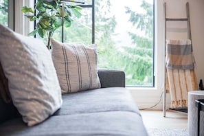 A grey sofa with striped pillows, a plant, and a ladder against a window with a view of greenery. - Gorgeous 2+Loft | Creekside | Free Parking (Whistler)