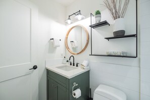 A modern bathroom with a white sink, a round mirror, and a toilet. There are shelves above the mirror holding toiletries and a plant. - Gorgeous 2+Loft | Creekside | Free Parking (Whistler)