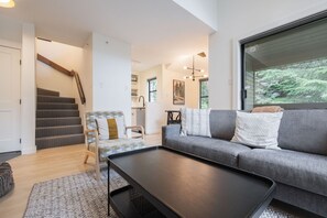 A modern living room with a grey sofa, a patterned armchair, a black coffee table, and a staircase leading to a kitchen area. - Gorgeous 2+Loft | Creekside | Free Parking (Whistler)