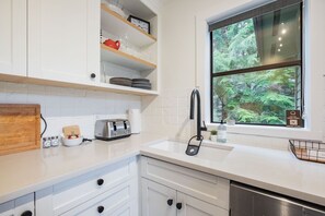 Une cuisine moderne avec des meubles blancs, un lave-vaisselle en acier inoxydable, un grille-pain et une fenêtre donnant sur des arbres.