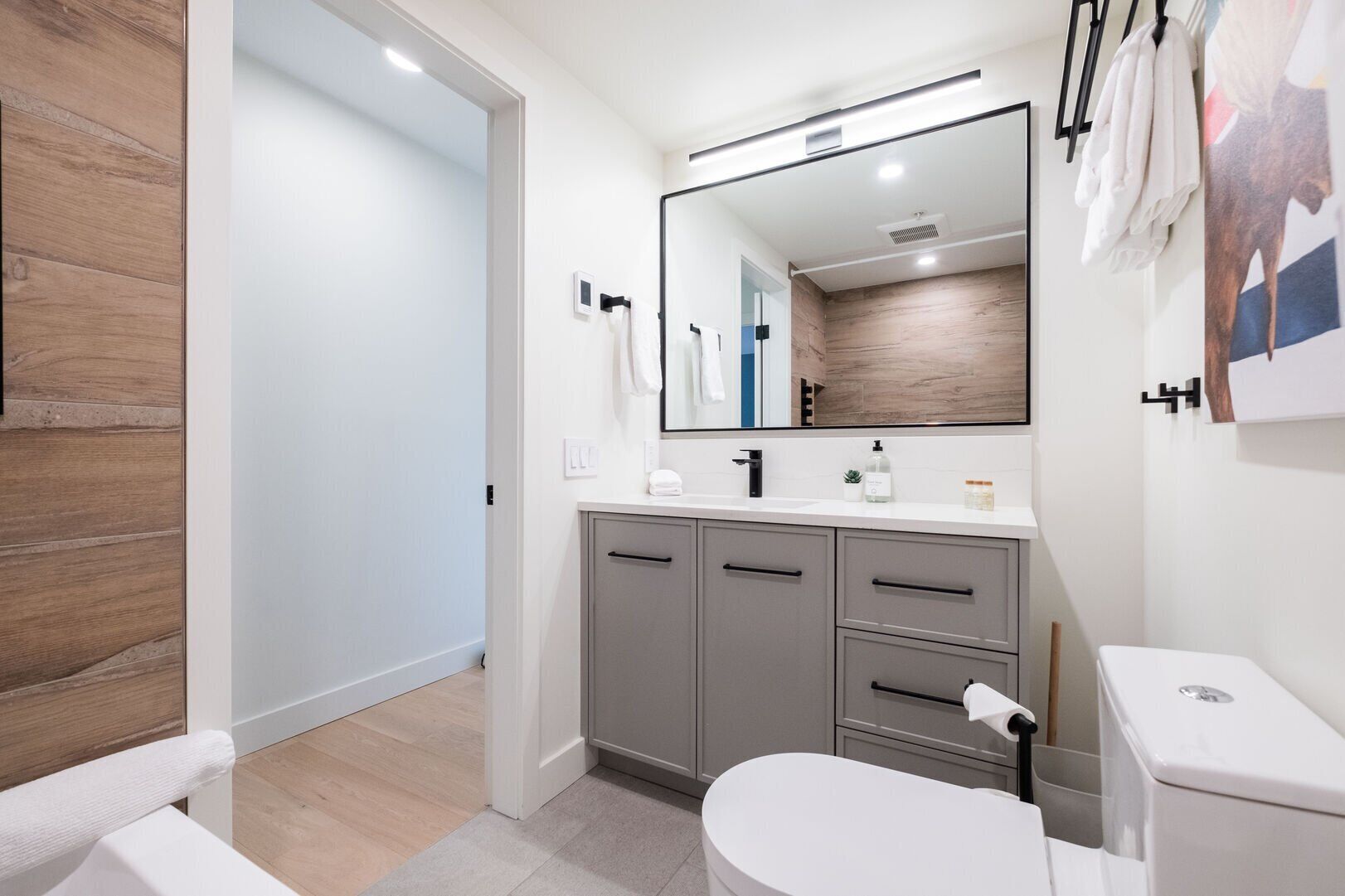 Ein modernes Badezimmer mit einer weißen Toilette, grauen Schränken, einem großen Spiegel und einer Wand mit Holzverkleidung.