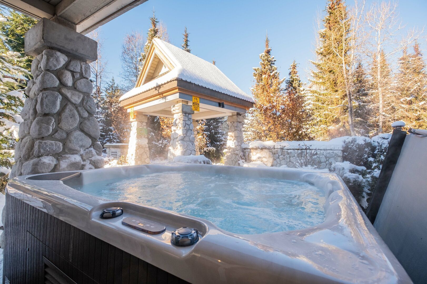 Un bain à remous doté d’un mur en pierre et d’une terrasse en bois, entouré d’arbres couverts de neige et d’une structure en pierre en arrière-plan.