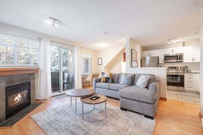 A modern living room with a fireplace, a grey sofa, a round coffee table, and a kitchen area with white cabinets and appliances. - Private Hot Tub | Pets | Free Parking | Location! (Whistler)