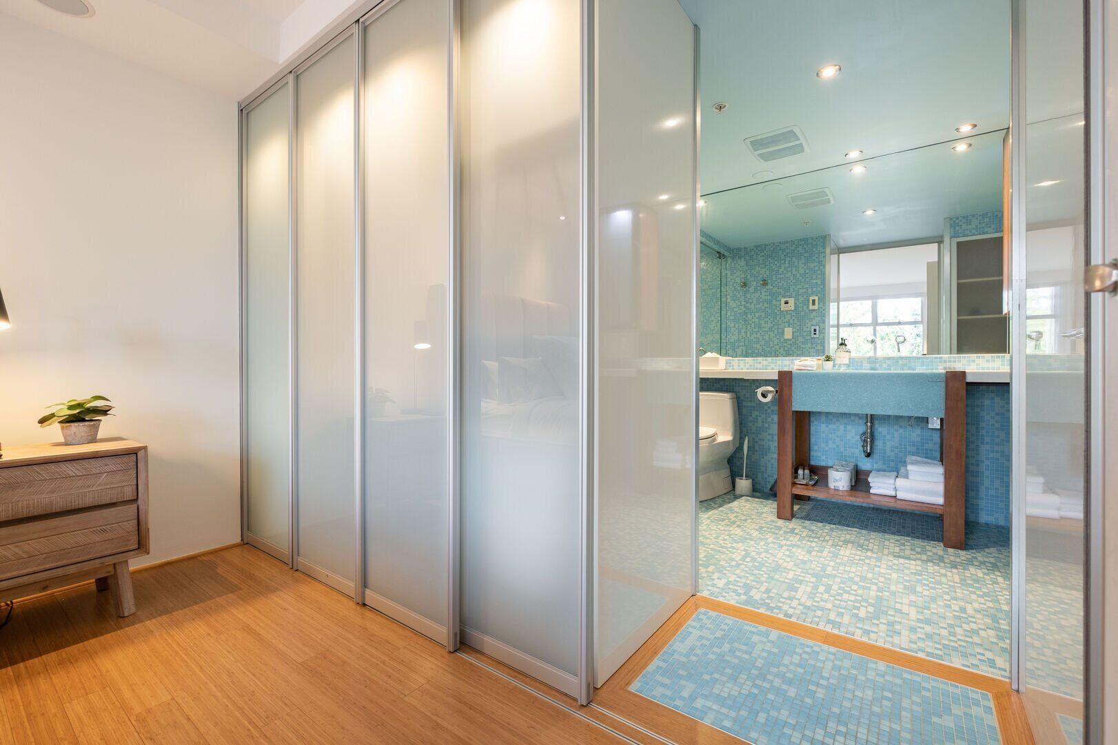 A modern bathroom with a glass partition, a wooden floor, a sink with a blue tiled backsplash, and a mirror above the sink.