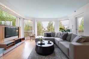 Ein modernes Wohnzimmer mit einem großen Flachbildfernseher, einer Couch, einem Couchtisch und großen Fenstern mit Blick auf Bäume.