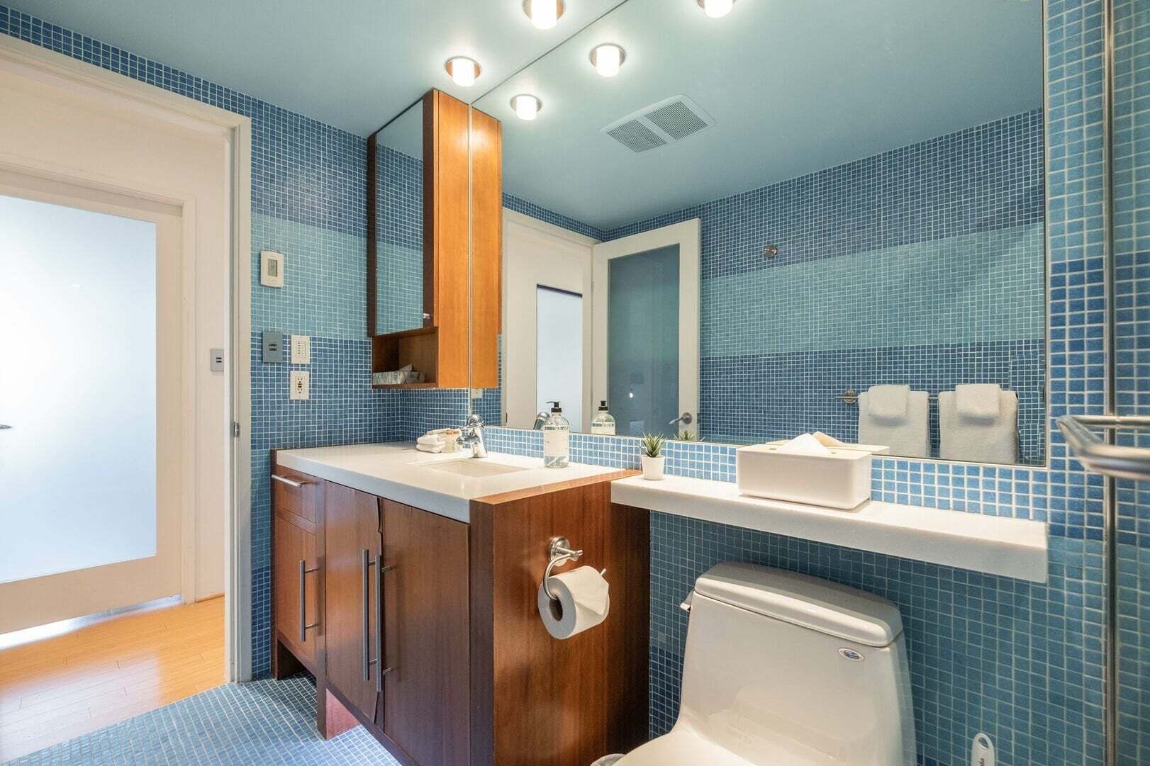 A bathroom with blue-tiled walls, a wooden vanity, a white sink, a mirror, and a toilet.