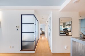 A modern interior with a hallway leading to a kitchen area, featuring a glass partition, a mounted mirror, and a wooden floor.