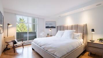 Ein modernes Schlafzimmer mit einem großen Bett, einem Sessel, einem Nachttisch, einer Stehlampe und einem Fenster mit Blick auf Bäume.