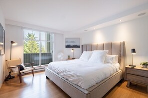 A modern bedroom with a large bed, a chair, a bedside table, a nightstand, a floor lamp, and a window with a view of trees.