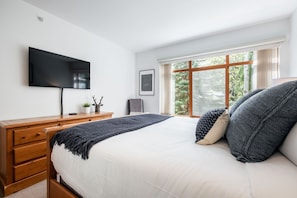 Ein Schlafzimmer mit einem Bett, einer Holzkommode, einem Fernseher und einem Fenster mit Blick auf Bäume.