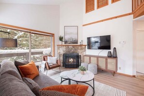 A living room with a fireplace, a sofa, a television, and a large window overlooking a snowy landscape. - 2+loft | Walk to Creekside Gondola | Pet friendly (Whistler)