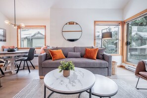 Ein modernes Wohnzimmer mit einem grauen Sofa, einem runden Couchtisch und einem Essbereich mit einem weißen Tisch und Stühlen.