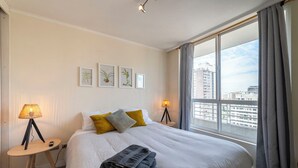 A hotel room with a large bed, two bedside lamps, a small table, and a view of the city. - San Isidro Ideal for Families 4 (Santiago)