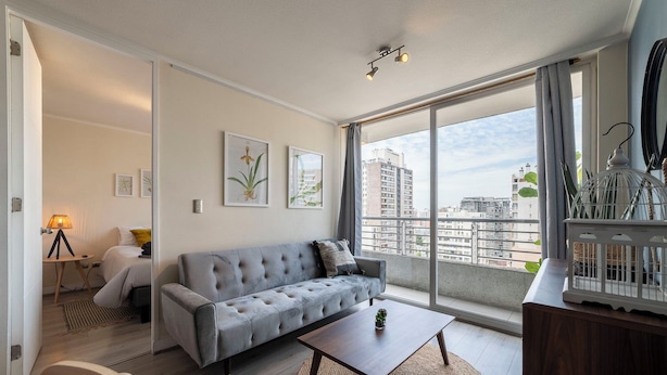 A modern living room with a grey sofa, a small table, and a view of the city through large windows. - San Isidro Ideal for Families 4 (Santiago)