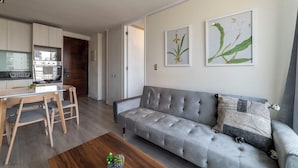 A modern living room with a grey sofa, wooden dining table, and chairs. There is a kitchen area with a microwave and oven. - San Isidro Ideal for Families 4 (Santiago)