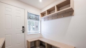 Une pièce avec une porte blanche, une fenêtre avec des stores, un banc et un porte-manteau fixé au mur.