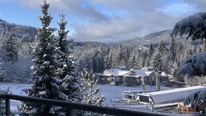 A snow-covered landscape with a building complex and a snow-laden tree in the foreground. - Heated pool, hot tub & gym | Ski-in/out Retreat (Whistler)