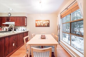 Une cuisine avec des meubles de rangement en bois, une table à manger avec des chaises, un tableau accroché au mur et une fenêtre donnant sur un paysage enneigé.
