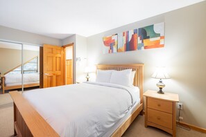 A bedroom with a wooden bed, bedside lamps, a wooden nightstand, and a large abstract painting on the wall. - BBQ | Private garage | Walk to Golfing & Lost Lake (Whistler)