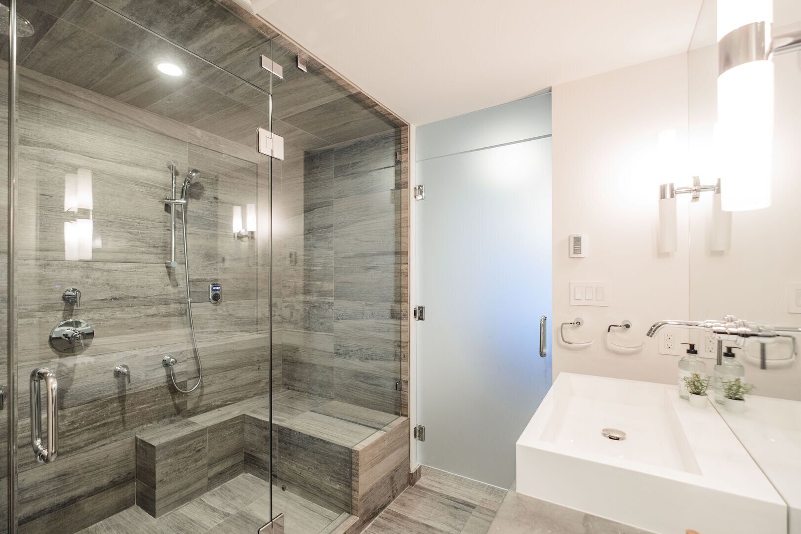 A modern bathroom with a glass-enclosed shower, a white sink with a mirror, and a light blue door.