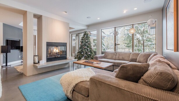 A modern living room with a fireplace, a Christmas tree, and a large window with a view of trees. - Gorgeous townhouse | Private hot tub | BBQ (Whistler)