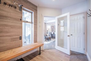 A modern interior with a wooden bench, a white door, and a view into a kitchen area. - Gorgeous townhouse | Private hot tub | BBQ (Whistler)
