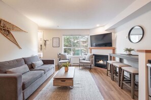 A living room with a sofa, armchairs, a coffee table, a fireplace, a TV, and a large window. - AC in bedrooms | Private hot tub | Pets | Location (Whistler)