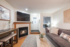 A living room with a fireplace, a sofa, a coffee table, a TV, and a door leading to the outside. - Beautiful 2BDR townhouse | Private hot tub | Pets (Whistler)